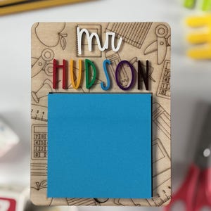 Pu&ograve; includere: Una lavagna in legno con il nome "mr HUDSON" in lettere 3D colorate. La lavagna ha un blocco note adesivo blu e uno sfondo di illustrazioni a tema scolastico. Matite e giocattoli colorati sullo sfondo.