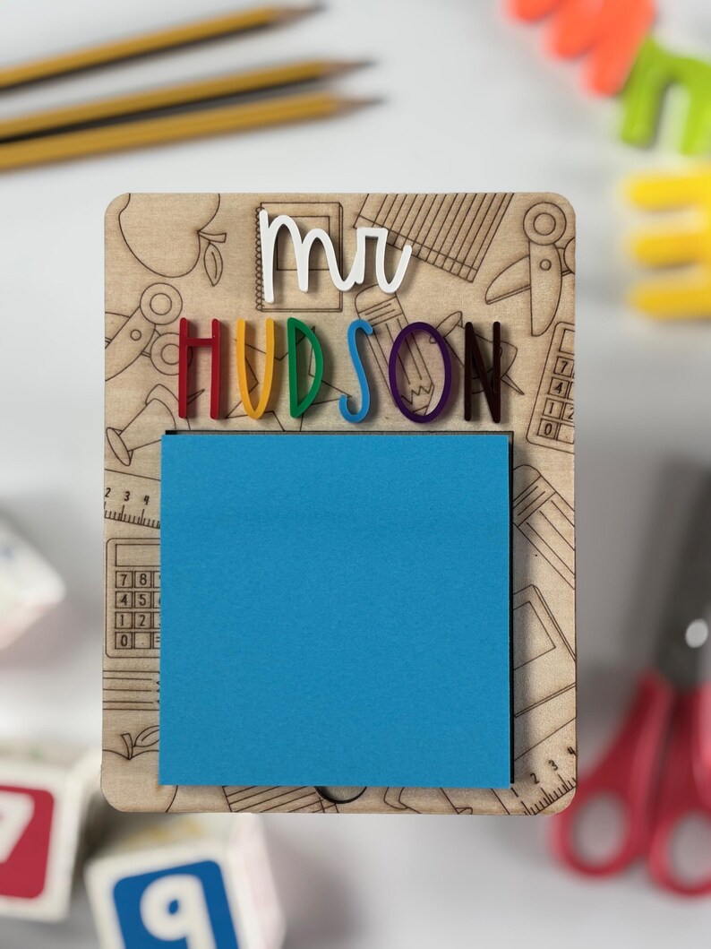 Pu&ograve; includere: Una lavagna in legno con il nome "mr HUDSON" in lettere 3D colorate. La lavagna ha un blocco note adesivo blu e uno sfondo di illustrazioni a tema scolastico. Matite e giocattoli colorati sullo sfondo.