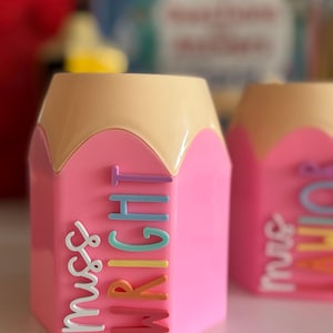 May include: A pink pencil-shaped desk organizer with a tan top. The side features the text "miss WRIGHT" in raised, multi-colored letters including white, light purple, light blue, teal, yellow, orange, and dark pink, arranged vertically.