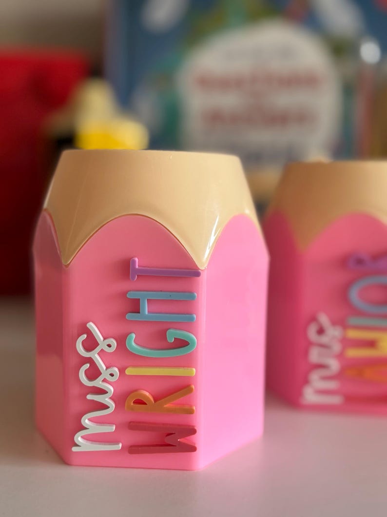 May include: A pink pencil-shaped desk organizer with a tan top. The side features the text "miss WRIGHT" in raised, multi-colored letters including white, light purple, light blue, teal, yellow, orange, and dark pink, arranged vertically.