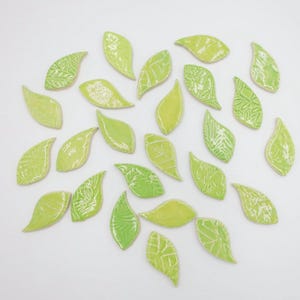 May include: A collection of twenty-five small, leaf-shaped ceramic tiles in various shades of lime and light green. Each piece features a glossy finish and unique embossed vein patterns, scattered across a plain white surface.