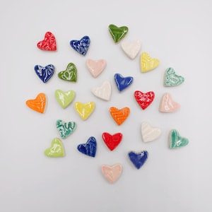 May include: A collection of colorful ceramic heart-shaped decorations in various colors, including red, blue, green, yellow, orange, and pink. Each heart has a unique textured design. The hearts are scattered on a white surface.