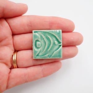 May include: A small, square, turquoise ceramic tile with a raised relief design of a stylized fish. The tile is held in a hand, showcasing the intricate details of the aquatic motif. The tile is a decorative element.