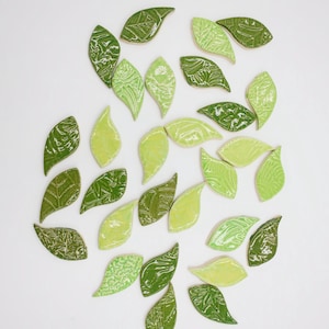 May include: Ceramic leaf tiles in shades of green, arranged on a white background. The leaves feature textured patterns and glossy finishes. Ideal for decorative purposes, these tiles offer a natural aesthetic.