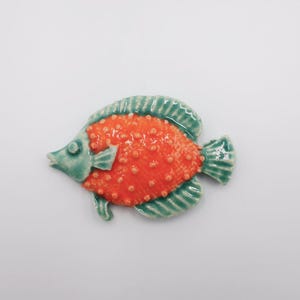 Peut inclure: Une figurine de poisson en céramique avec un corps orange et des nageoires turquoise. Le poisson a une surface texturée et est posé sur un fond blanc. La figurine est probablement un objet décoratif.