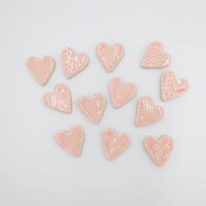 May include: A collection of eleven pink ceramic heart-shaped tiles with various embossed patterns, including chevrons and dots. The tiles are scattered on a white surface, creating a decorative arrangement. Each heart is approximately 1 inch in size.