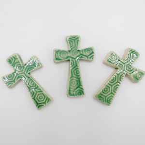 May include: Three ceramic crosses with a green geometric pattern on a white background. The crosses are in varying orientations, showcasing their design. The crosses have a glossy finish and a detailed, textured surface.