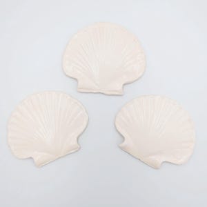 May include: Three off-white seashell-shaped decorative items. Each shell has a detailed, ridged texture, and a curved, fan-like shape. The shells are arranged on a plain white background, suggesting a coastal or nautical theme.