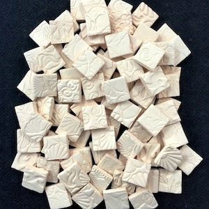 Op de afbeelding: Een stapel crèmekleurige keramische tegels met verschillende geëmbosseerde patronen. De tegels zijn vierkant en hebben een licht getextureerd oppervlak.