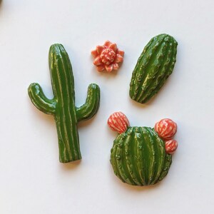 Op de afbeelding: Vier keramische cactus-figuurtjes in groen en roze. De figuurtjes zijn in verschillende vormen en maten, waaronder een hoge cactus met een enkele stengel, een ronde cactus met twee kleine roze bloemen en een lange, dunne cactus.