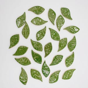 Op de afbeelding: Een verzameling glanzende, groene keramische tegels in de vorm van bladeren met gedetailleerde aderpatronen. De bladeren zijn op een witte achtergrond gerangschikt, wat een decoratieve en natuurlijke uitstraling creëert. De tegels zijn waarschijnlijk voor woondecoratie of knutselprojecten.