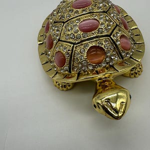 May include: A gold turtle figurine with pink and clear rhinestones. The turtle's shell is decorated with a geometric pattern.
