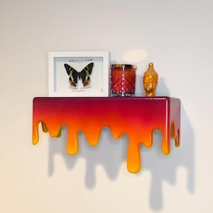 May include: A shelf with a dripping paint effect in shades of yellow, orange, and red. The shelf is decorated with a framed butterfly specimen, a glass candle holder, and a gold Buddha statue.
