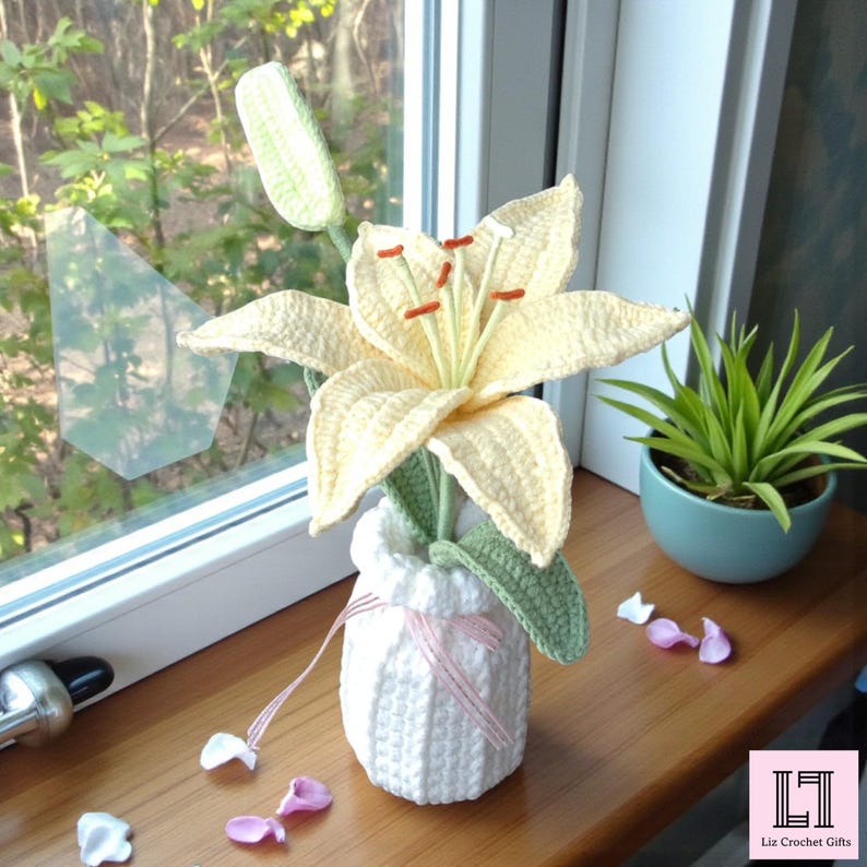May include: A crocheted pale yellow lily flower with orange stamens and green leaves, displayed in a white crocheted vase. The vase is tied with a thin pink ribbon and sits alongside scattered white and pink petals on a wooden surface.