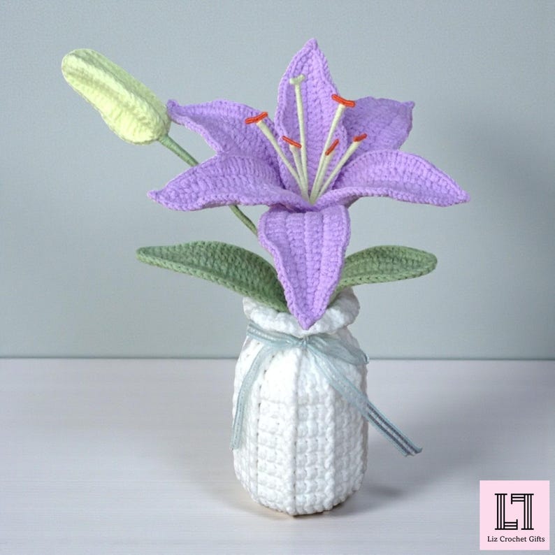 May include: A handcrafted crochet lily flower with light purple petals, orange-tipped stamens, and a pale green bud. The flower is arranged in a white crocheted vase tied with a thin, light blue ribbon. The text Liz Crochet Gifts is visible.