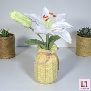 May include: A crocheted white lily flower with orange stamens and a light green bud, set in a textured yellow crocheted vase tied with a thin pink ribbon. The flower features green crocheted leaves and stems, creating a soft, handcrafted appearance.