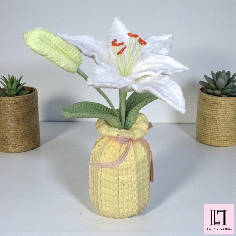 May include: A crocheted white lily flower with orange stamens and a light green bud, set in a textured yellow crocheted vase tied with a thin pink ribbon. The flower features green crocheted leaves and stems, creating a soft, handcrafted appearance.