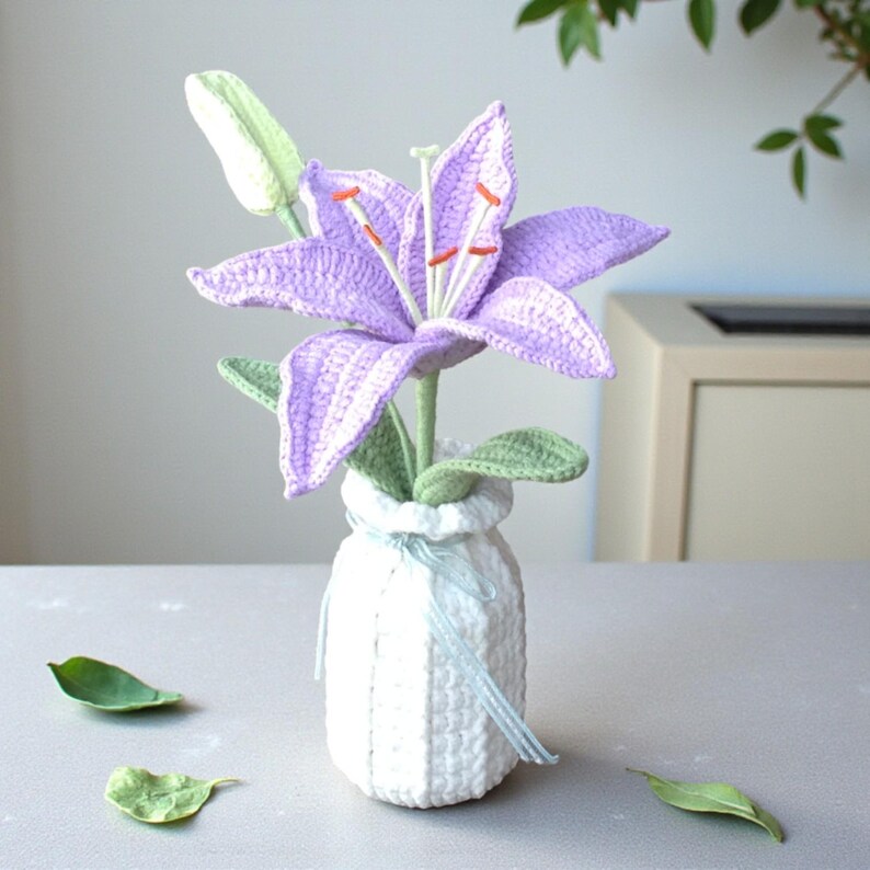 May include: A crocheted purple lily flower with orange-tipped stamens and a light green bud, set in a white crocheted vase. The vase is tied with a thin, light blue ribbon. The lily has textured green leaves attached to its stem.