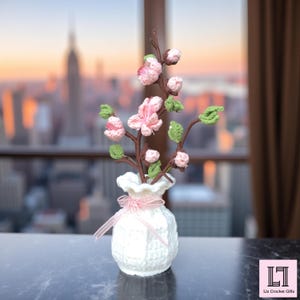 May include: A handcrafted crochet floral arrangement featuring pink blossoms, green leaves, and brown stems, displayed in a white crochet vase tied with a pink ribbon. The vase is approximately 5 inches tall. The background shows a cityscape.