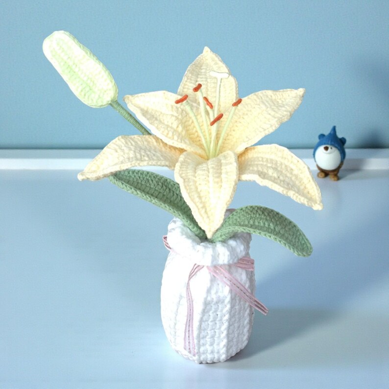 May include: A crocheted cream-colored lily flower with orange stamens, a green stem, and green leaves, displayed in a white crocheted vase tied with a thin pink ribbon. A small blue and white gnome figurine stands to the right of the vase.