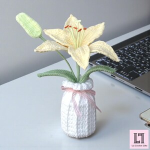 May include: A crocheted pale yellow lily flower with orange stamens and a light green bud, set in a white crocheted vase tied with a thin pink ribbon. The base of the vase features a small pink square logo with the text Liz Crochet Gifts.