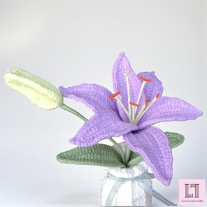 May include: A crocheted lavender lily flower with a pale green bud and green leaves, set in a white textured vase tied with a light blue ribbon. The flower features prominent white stamens with orange tips. The text Liz Crochet Gifts is visible.