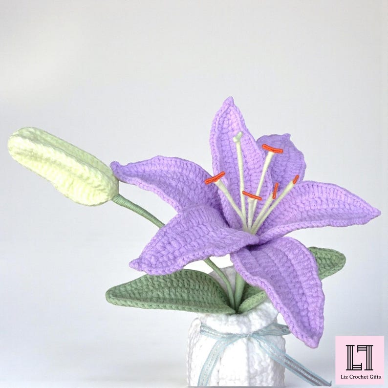 May include: A crocheted lavender lily flower with a pale green bud and green leaves, set in a white textured vase tied with a light blue ribbon. The flower features prominent white stamens with orange tips. The text Liz Crochet Gifts is visible.