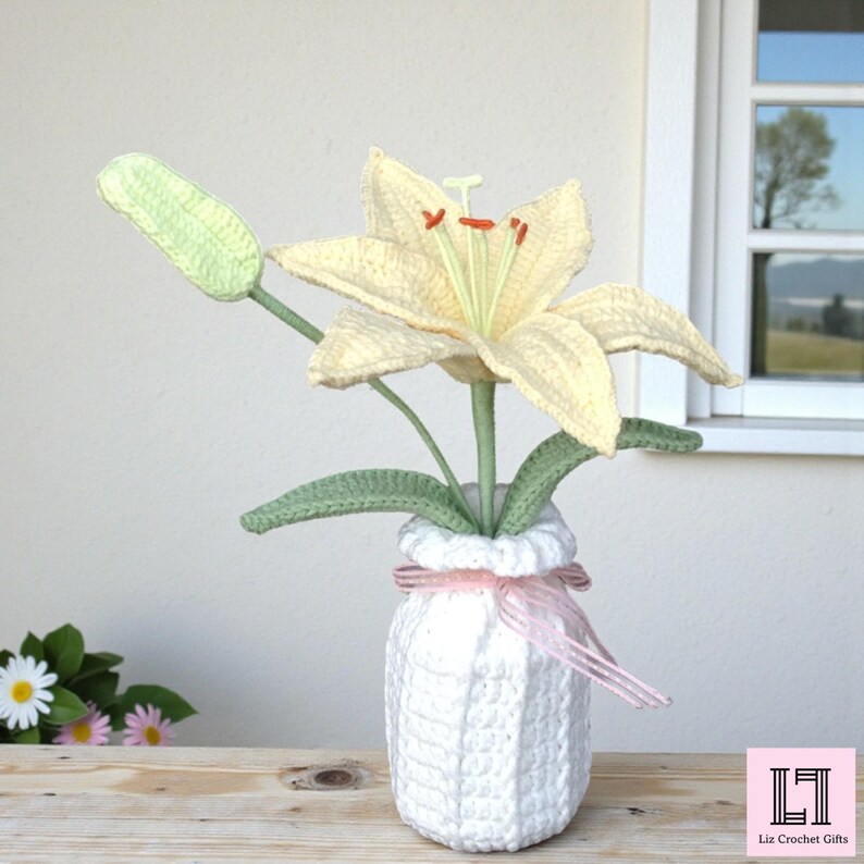 May include: A crocheted pale yellow lily flower with a green bud and leaves, displayed in a white crocheted vase tied with a thin pink ribbon. The flower features prominent orange stamens and a light green pistil, all crafted with detailed yarn stitches.