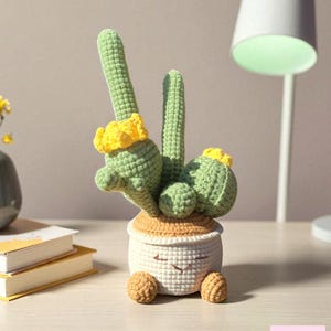 May include: A crocheted cactus plant with light green stems and bright yellow flowers, potted in a white, round, crocheted container featuring a stitched smiling face, tan top rim, and two small tan feet at the base, with the text Liz Crochet Gifts.