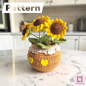 Könnte beinhalten: Eine gehäkelte Sonnenblumenpflanze in einem beigen Topf mit gelben Herzakzenten und weißer Spitzenborte. Die Sonnenblumen haben braune Mitten, leuchtend gelbe Blütenblätter und grüne Blätter. Das Wort Pattern steht oben links, das Logo von Liz Crochet Gifts unten rechts.