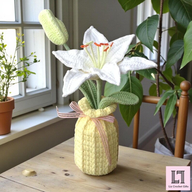 May include: A crocheted white lily flower with orange stamens and a green bud, displayed in a textured pale yellow crocheted vase. The vase is decorated with a thin pink and white striped ribbon tied in a bow around the neck.