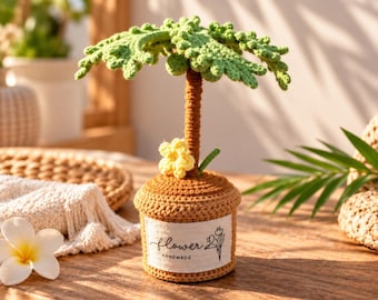 May include: A crocheted palm tree in a brown pot with a white label reading flower HANDMADE and a small yellow flower at the base. The tree has a textured brown trunk and green crocheted leaves. The item is crafted from yarn.