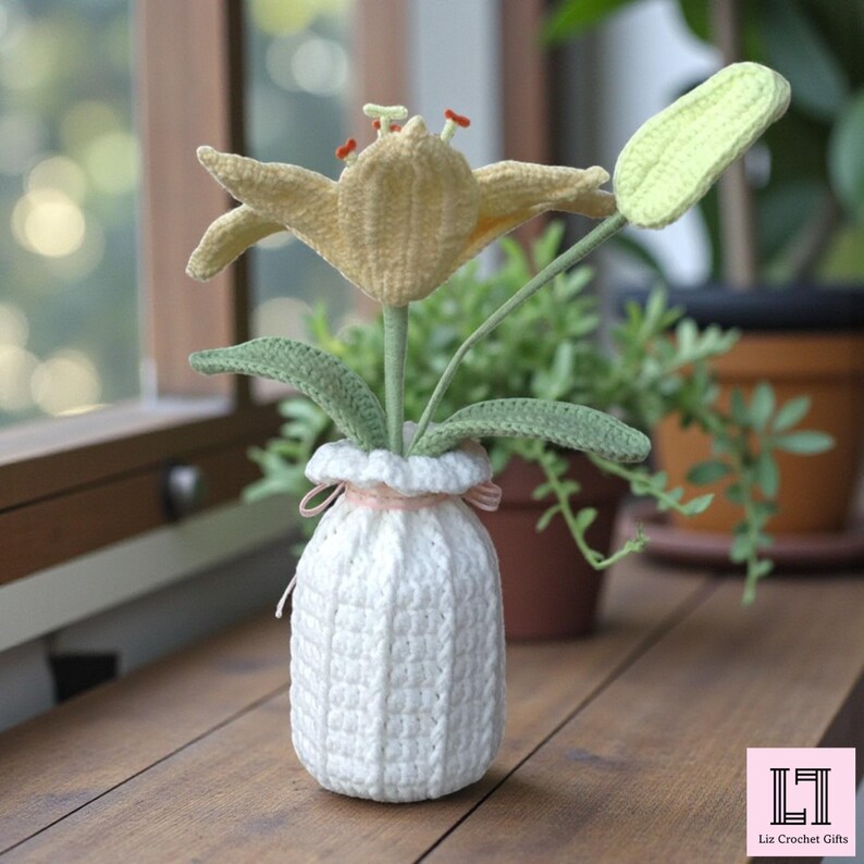 May include: A crocheted yellow lily flower with green leaves and a closed bud, arranged in a white crocheted vase tied with a thin pink ribbon. The vase features a textured vertical stitch pattern. A logo with the text Liz Crochet Gifts is visible.