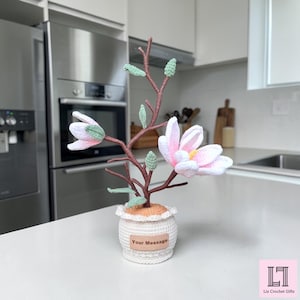 May include: A handcrafted crochet magnolia tree featuring a brown stem, light green leaves, and pale pink flowers with yellow centers. The tree is potted in a white crocheted base with a tan rectangular label that reads Your Message.