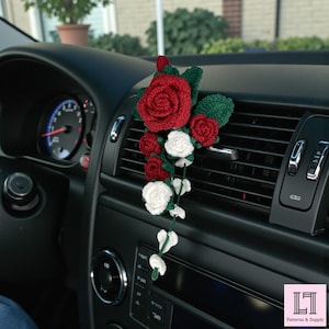 May include: Crocheted rose car air vent decoration featuring red and white roses with green leaves. The handmade floral accessory adds a touch of color and charm to a vehicle's interior. The roses are attached to a green vine.