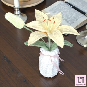 May include: A crocheted pale yellow lily flower with orange stamens and green leaves, set in a white crocheted vase tied with a thin pink ribbon. The flower has one closed bud and one open bloom with six petals, all crafted from yarn.