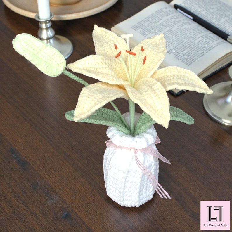 May include: A crocheted pale yellow lily flower with orange stamens and green leaves, set in a white crocheted vase tied with a thin pink ribbon. The flower has one closed bud and one open bloom with six petals, all crafted from yarn.