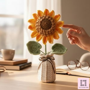 May include: A handcrafted crochet sunflower with yellow and orange petals and a brown center. The sunflower sits in a small, off-white vase tied with a brown ribbon. Green leaves and stem complete the floral arrangement.