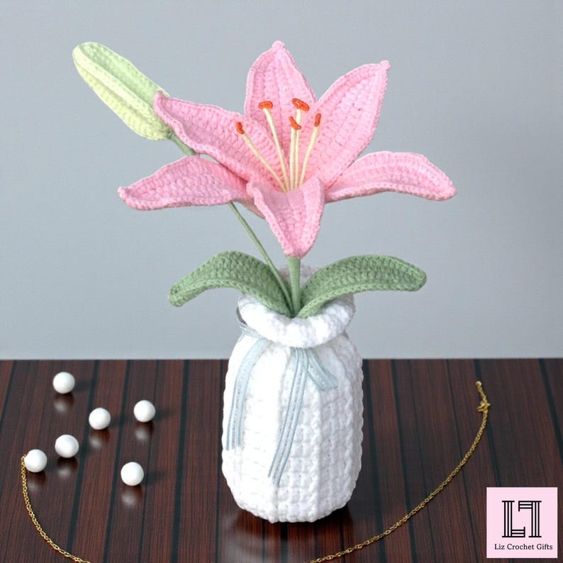 May include: A crocheted pink lily flower with a light green bud and leaves, arranged in a white textured crocheted vase tied with a light blue ribbon. The flower features prominent orange-tipped stamens. The text Liz Crochet Gifts is visible.