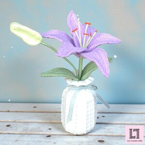 May include: A crocheted purple lily flower with a light green bud and green leaves, arranged in a white crocheted vase tied with a light blue ribbon. The flower features orange-tipped stamens. A logo with the text Liz Crochet Gifts is visible.