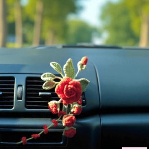 May include: A red crocheted rose and strawberry car air freshener. The handmade floral decoration features a large red rose, green leaves, and several red strawberries. The air freshener is displayed on a car dashboard.