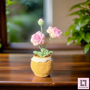 May include: A crocheted flower arrangement in a yellow pot with a white lace trim. The flowers are pink and white and have green leaves.