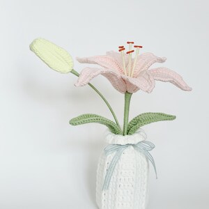 May include: A handcrafted crochet lily flower with pale pink petals, a light green bud, and green leaves, standing in a white crocheted vase. The vase is decorated with a thin, light blue and white striped ribbon tied in a small bow.