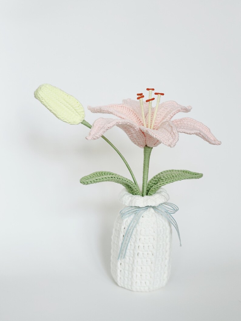 May include: A handcrafted crochet lily flower with pale pink petals, a light green bud, and green leaves, standing in a white crocheted vase. The vase is decorated with a thin, light blue and white striped ribbon tied in a small bow.