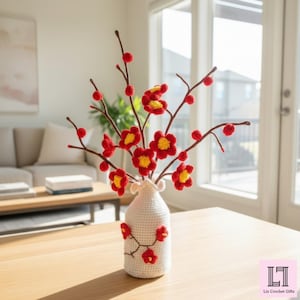 May include: A white crocheted vase with a ruffled top edge, decorated with small red and yellow crocheted flowers on the exterior. The vase holds several brown branches adorned with red and yellow crocheted blossoms and buds.