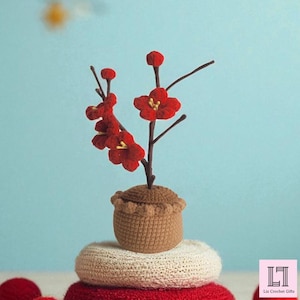 May include: A handcrafted crochet flower arrangement featuring vibrant red blossoms and brown stems, set in a small brown pot. The arrangement sits atop a stack of crocheted cushions in white and red. The background is a light blue.