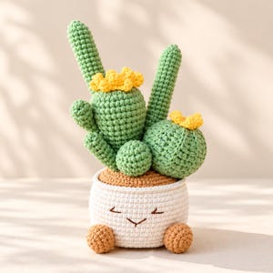May include: A crocheted cactus plant with green stems and yellow flowers, potted in a white crocheted container featuring a simple embroidered face and two small brown feet at the base, all crafted with visible yarn stitches.
