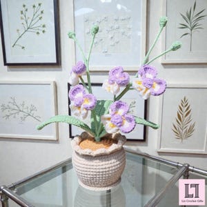 May include: A handcrafted crochet orchid plant featuring purple and white petals with yellow centers, green stems, and buds. The plant is potted in a textured, beige crocheted pot with a ruffled rim and a tan, soil-like crocheted top surface.