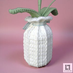 May include: A white crocheted jar cover with a textured, ribbed pattern and a ruffled top edge. A light blue ribbon is tied around the neck. Emerging from the top are several light green crocheted leaves. The text Liz Crochet Gifts is visible.