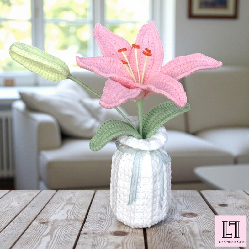 May include: A crocheted pink lily flower with green leaves and a closed bud, arranged in a white crocheted vase with a light blue ribbon. The vase features a textured grid pattern. A small pink square logo with the text Liz Crochet Gifts.